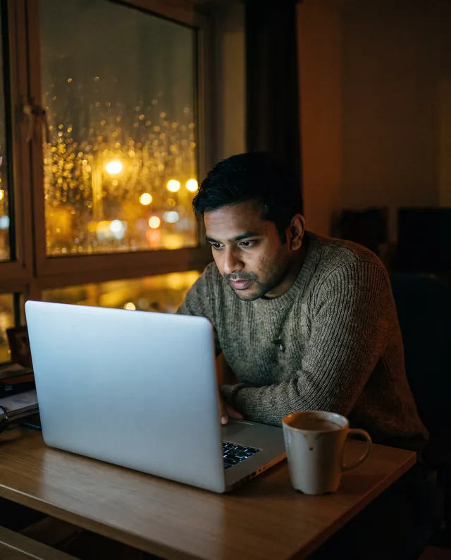 A man in a sweater reads a laptop screen late at night, with city lights blurred through a rain-spotted window behind him.