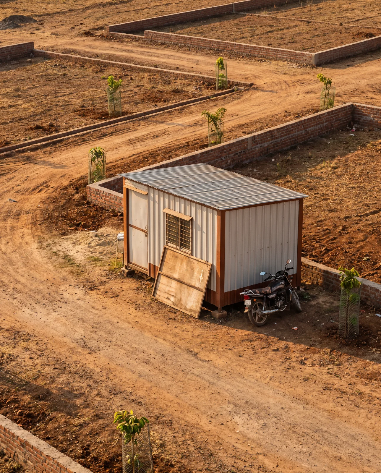 A tin-roofed site office and a parked motorcycle among brick plot walls and dirt roads in a new plotted colony