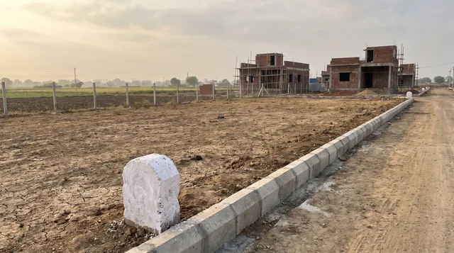 A whitewashed boundary stone stands at the corner of a bare, cracked plot beside a fresh concrete kerb and an unmade road, with brick houses under construction and open fields behind.