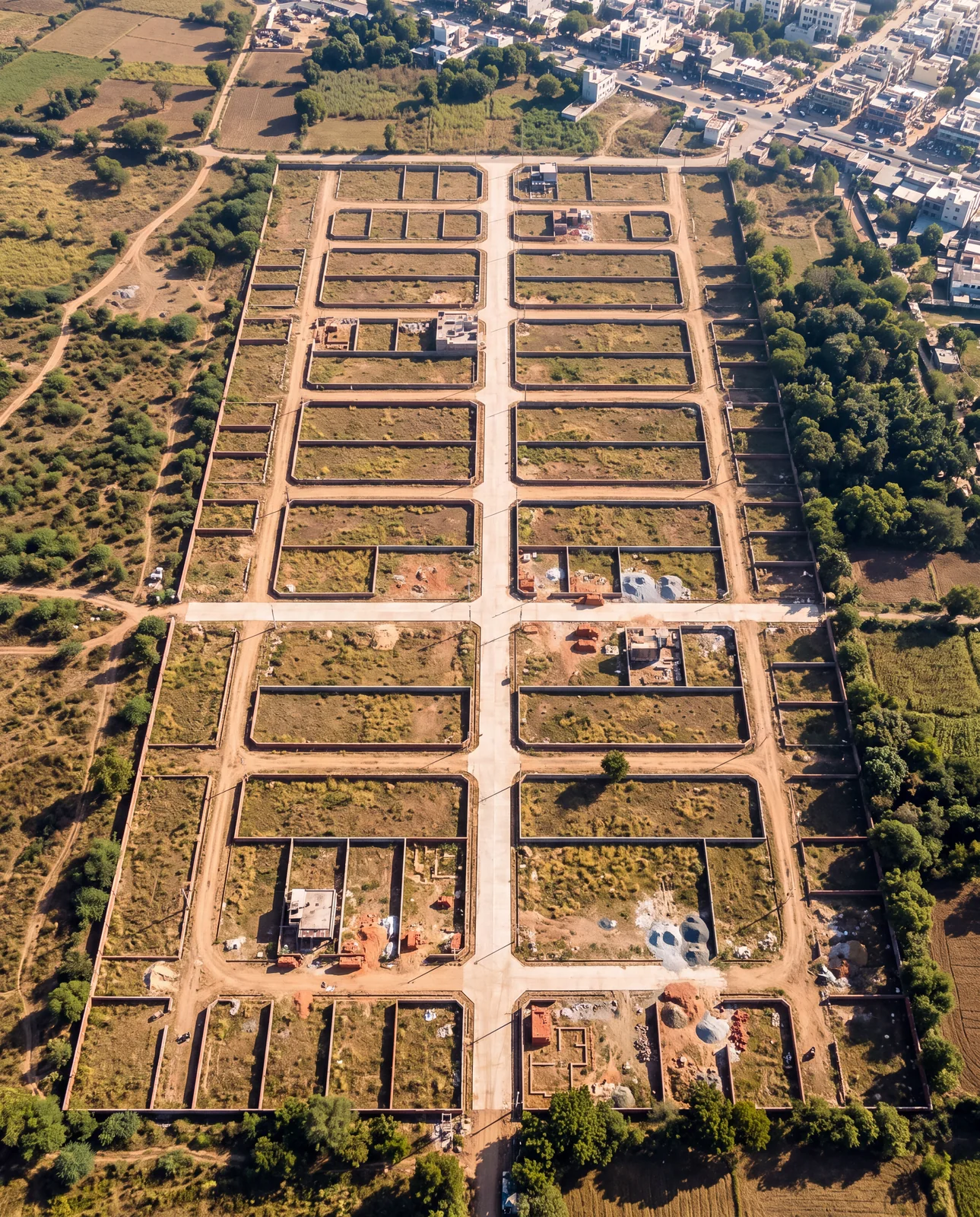 An aerial view of a plotted colony at the edge of a city, its compound walls and concrete roads laid out in a grid with almost no houses built yet.