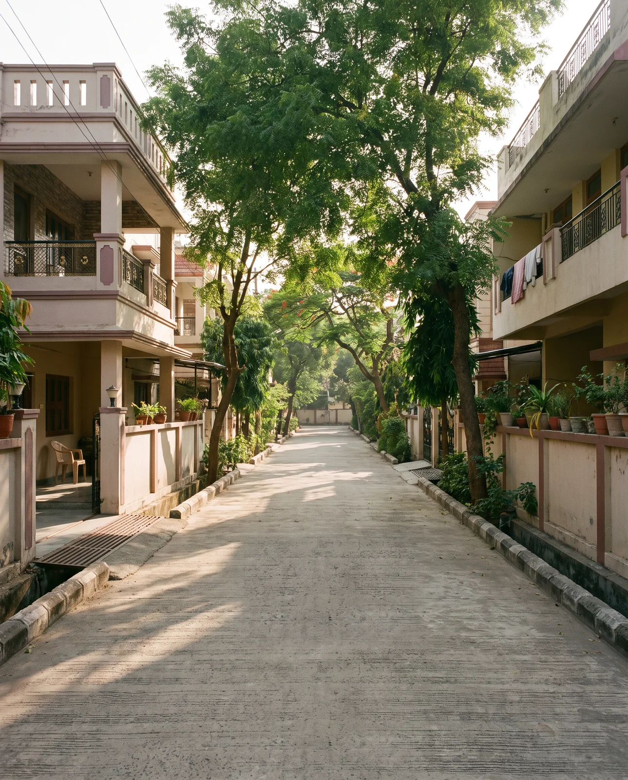 A quiet tree-lined colony street with a concrete road, kerbs, covered drains and houses with balconies on both sides