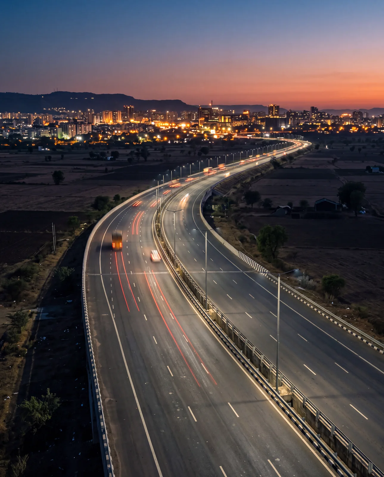 A lit expressway curving across dark fields towards a city skyline glowing at dusk under an orange sky.