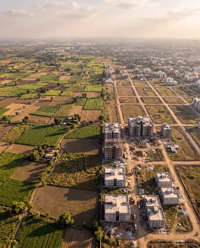 An aerial view of patchwork farmland on the left meeting a grid of new dirt roads, half-built apartment blocks and the city spreading to the horizon on the right.