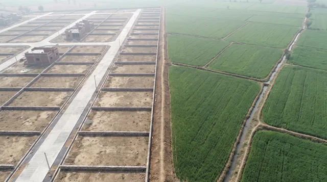 An aerial view of walled, empty plots with concrete roads on the left and green wheat fields with an irrigation channel on the right, divided by a raised earth bund.