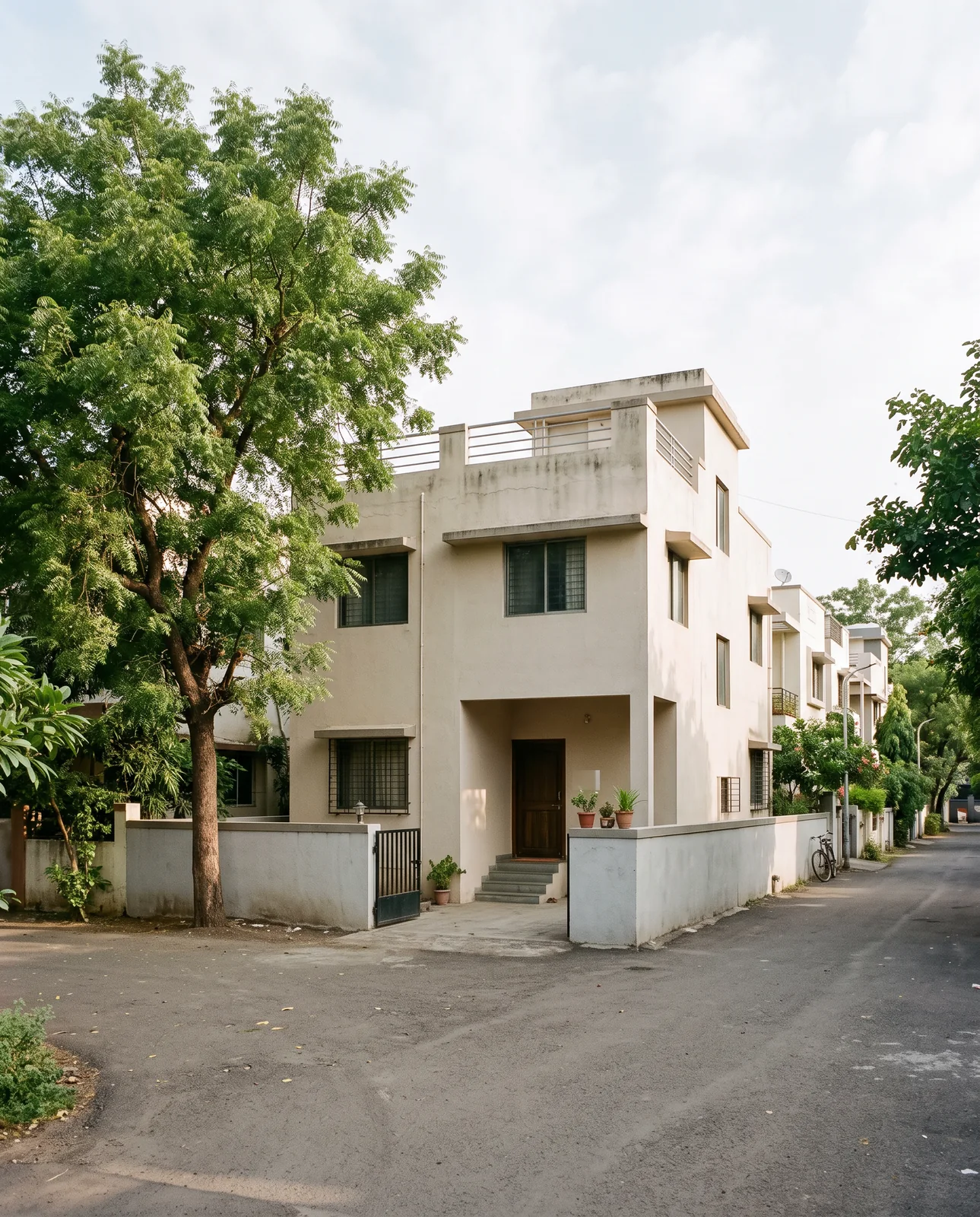 A two-storey cream house with a small gate and a neem tree at a quiet colony corner