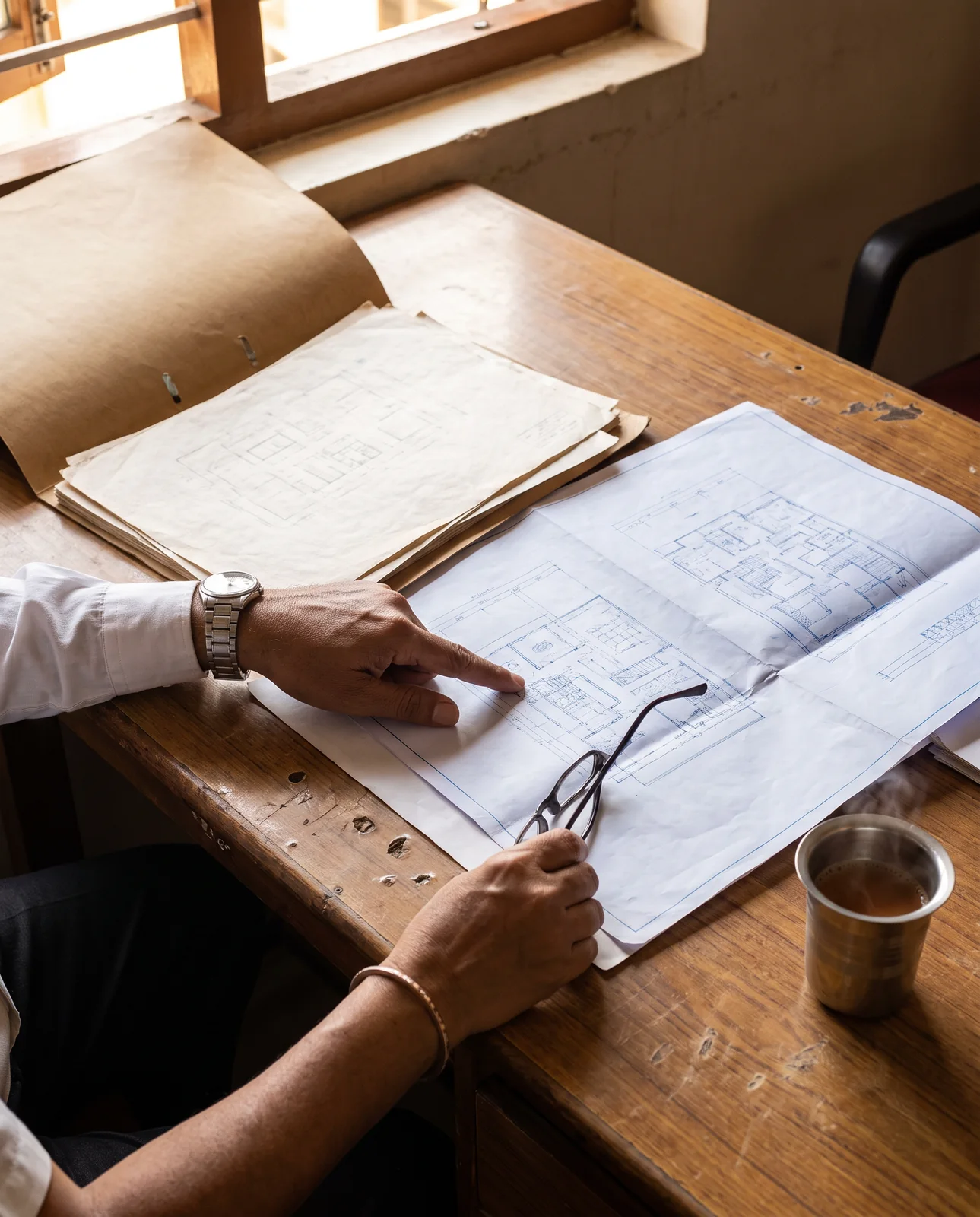 A hand points at a blueprint spread on a worn wooden desk, with a folder of older drawings and a steel glass of tea beside it