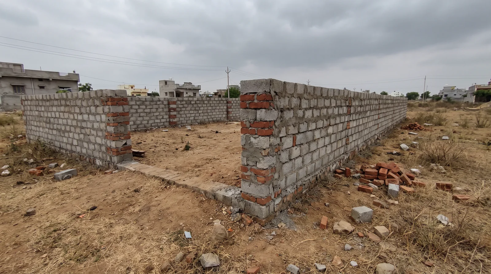 A half-built boundary wall of grey concrete blocks with red-brick corner pillars encloses a bare plot under a heavy grey sky, loose bricks stacked beside it.
