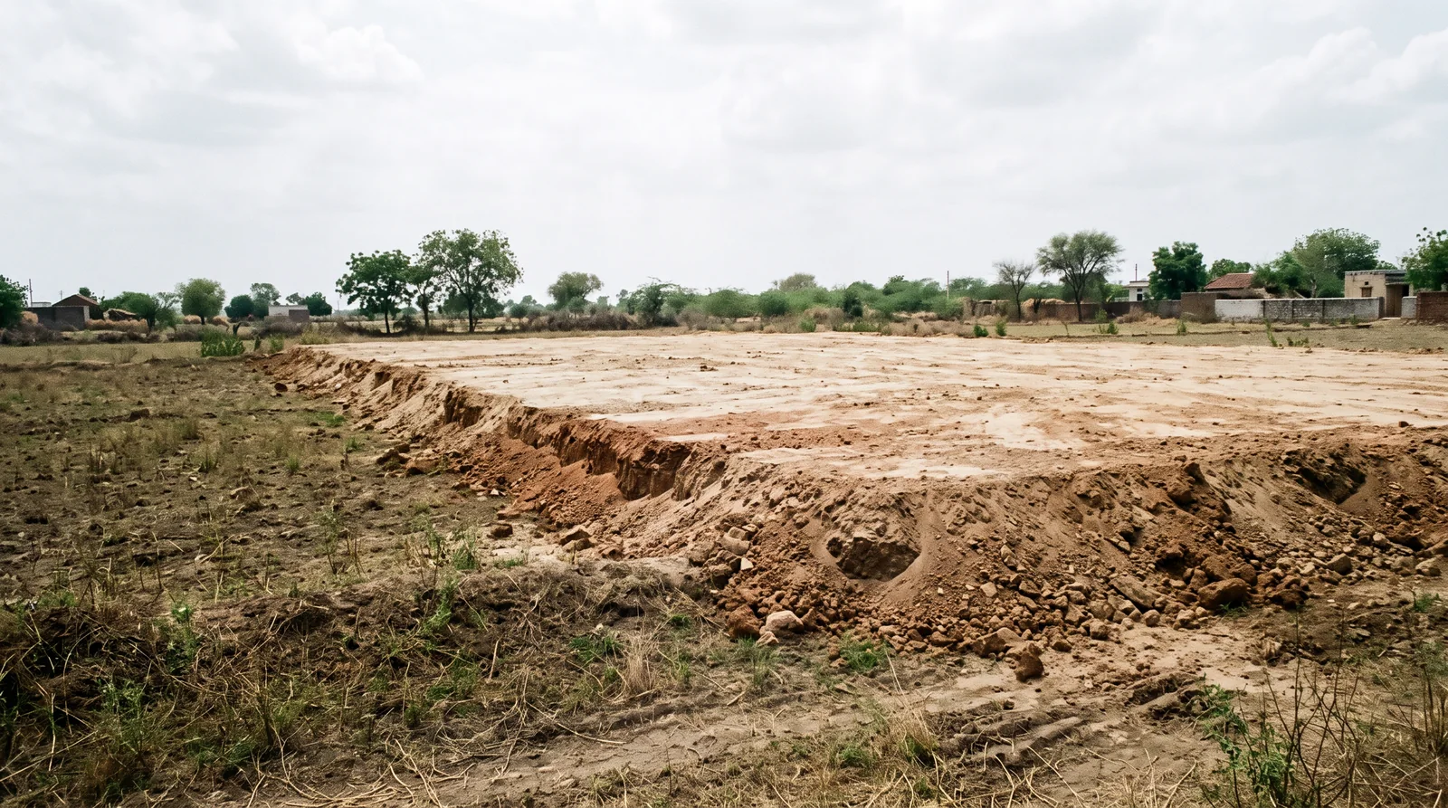 A raised platform of freshly dumped pale earth, its edge a rough step above the older, darker ground and scrub beside it, with trees and houses in the distance.