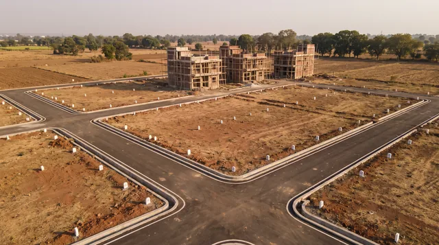 A freshly laid grid of tarmac roads with white kerbs and boundary stones marking empty plots, three brick houses under construction behind them and farmland beyond, seen from above in late-afternoon light.